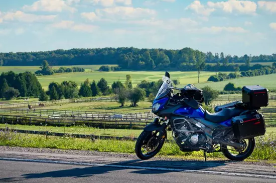 Une motocyclette bleue garée sur le côté d’une route rurale surplombant une ferme équestre.